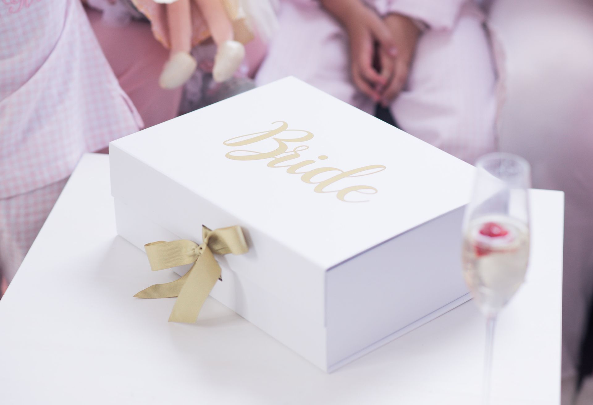 White bridal gift box with gold ribbon and “Bride” lettering on a table beside a champagne flute
