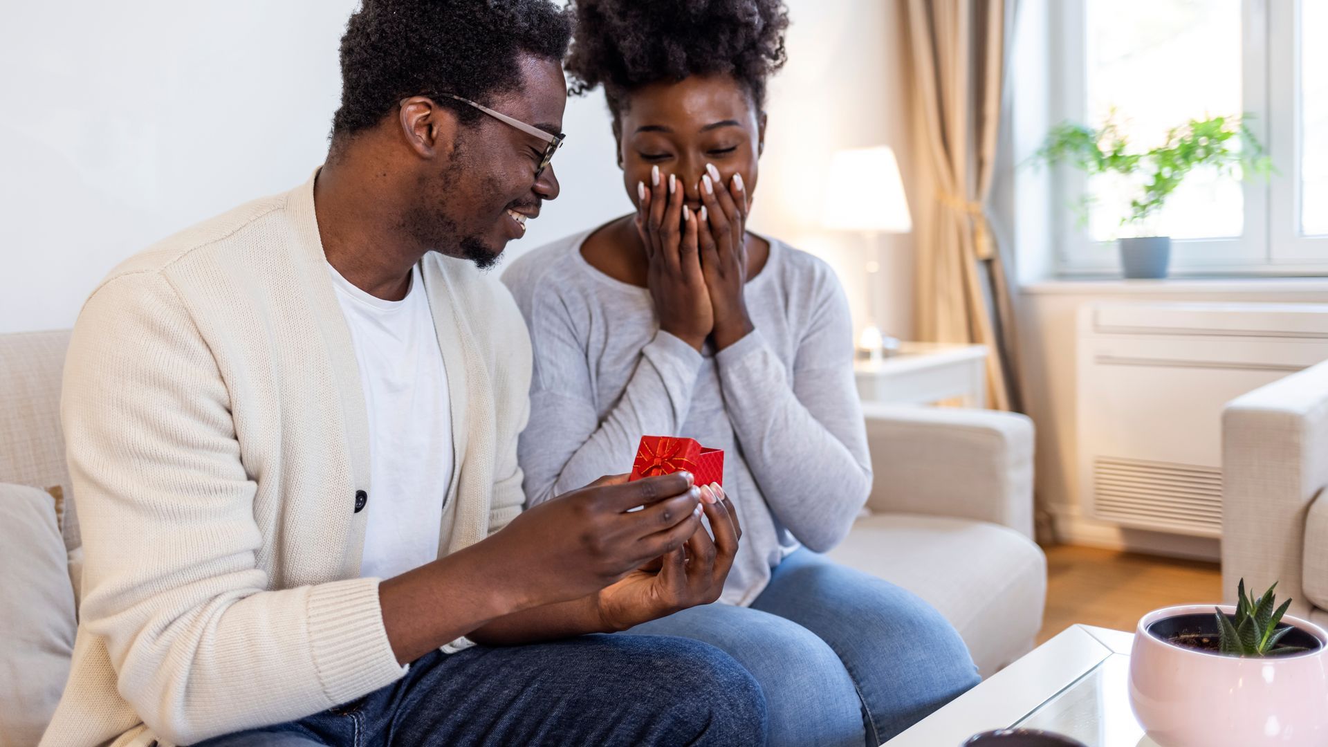Couple on a couch, smiling as one presents a small red gift box in a bright living room.
