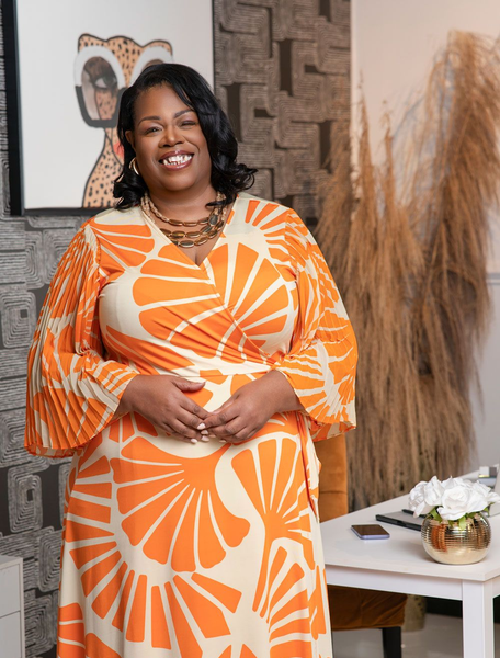 Woman in an orange patterned dress smiling indoors beside a white table and decorative vase.