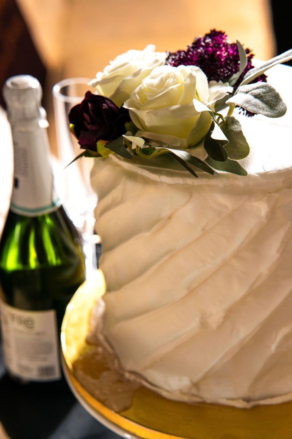 White frosted cake with flower topper beside a champagne bottle on a table