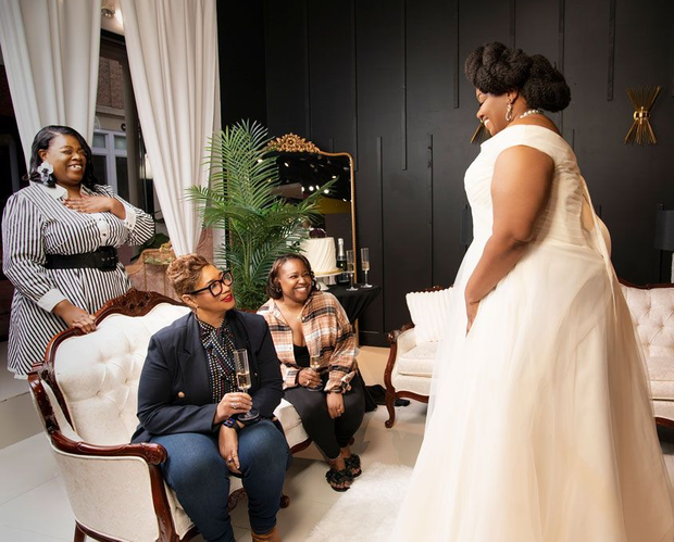 Bridal party in a living room, with a woman in a white dress facing seated guests.