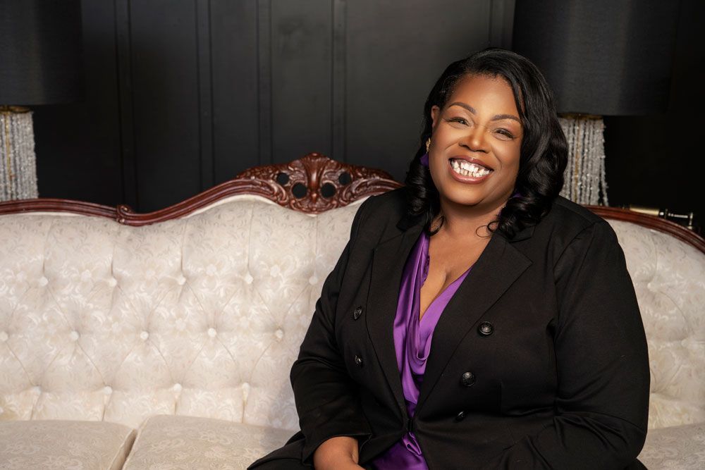 Smiling woman in a black blazer and purple blouse seated on a tufted white sofa