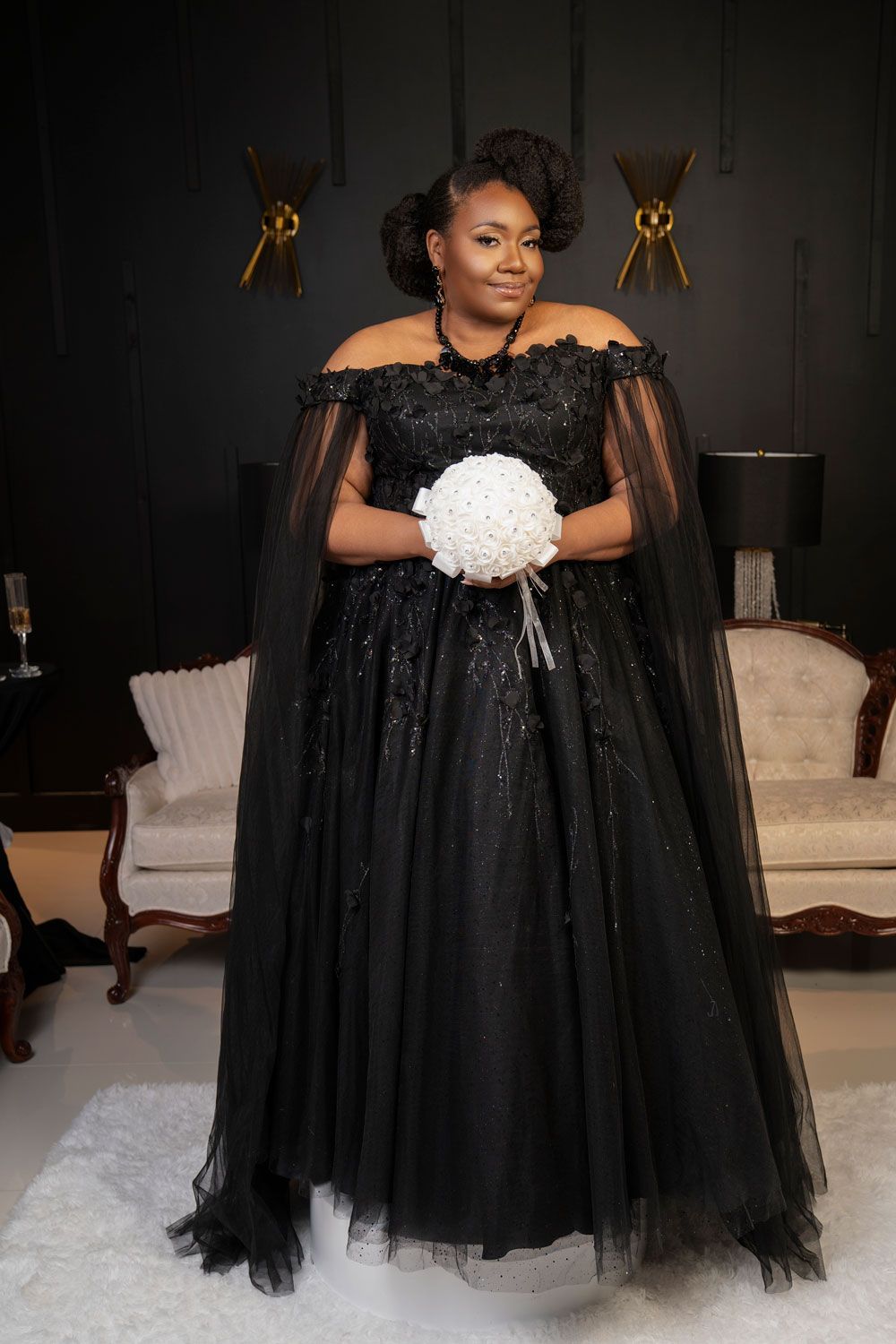 Woman in a black gown with a lace cape, holding a white bouquet, standing in an elegant room.