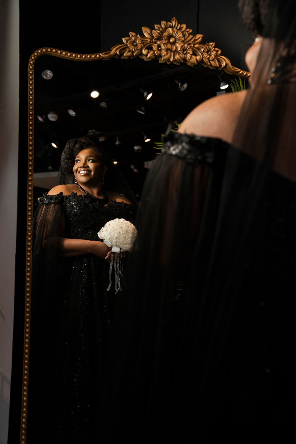 Bride in black dress holding white bouquet, smiling into a mirror with ornate gold frame.