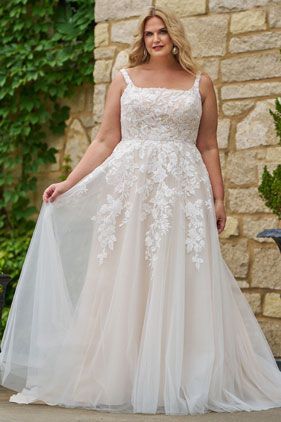 Woman in a white lace wedding dress standing against a stone wall, holding out the tulle skirt.