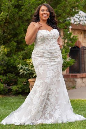 Woman in a white lace wedding gown posing outdoors in a garden, smiling.