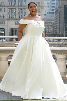 Woman in an off-shoulder white gown smiling on a rooftop with city buildings behind her