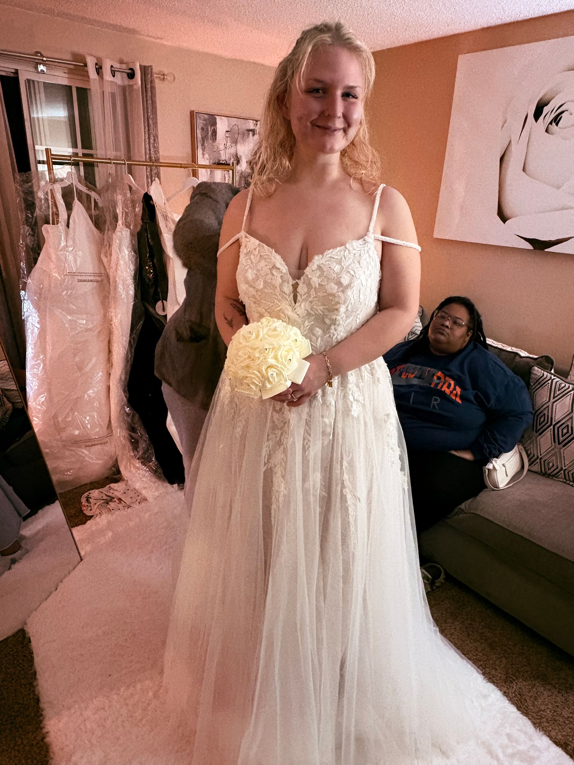 Bride in a white lace gown holding a bouquet, standing in a softly lit room with dresses in the background