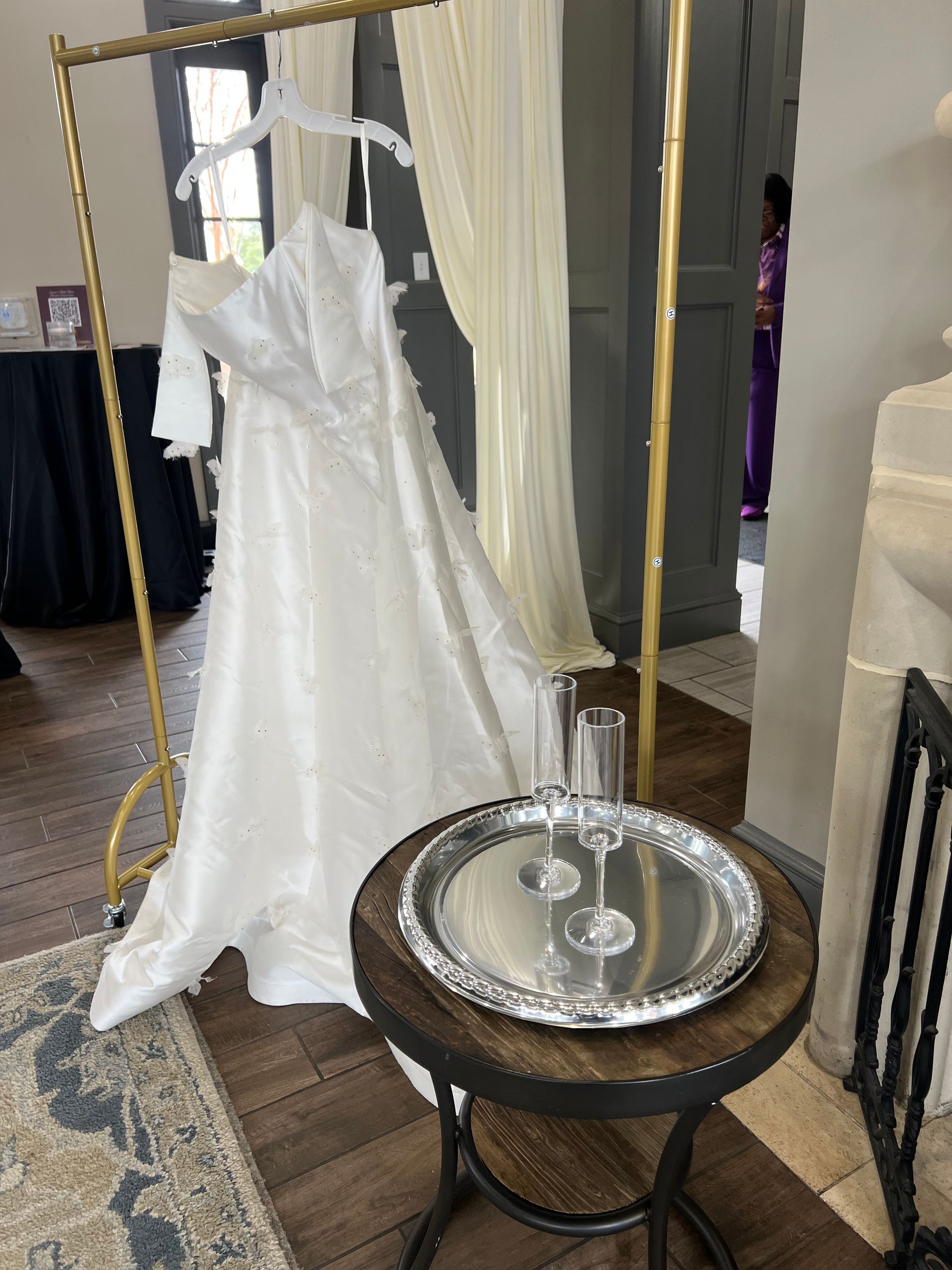 White wedding dress on a gold rack beside a small table with a silver tray and glasses in a fitting room.