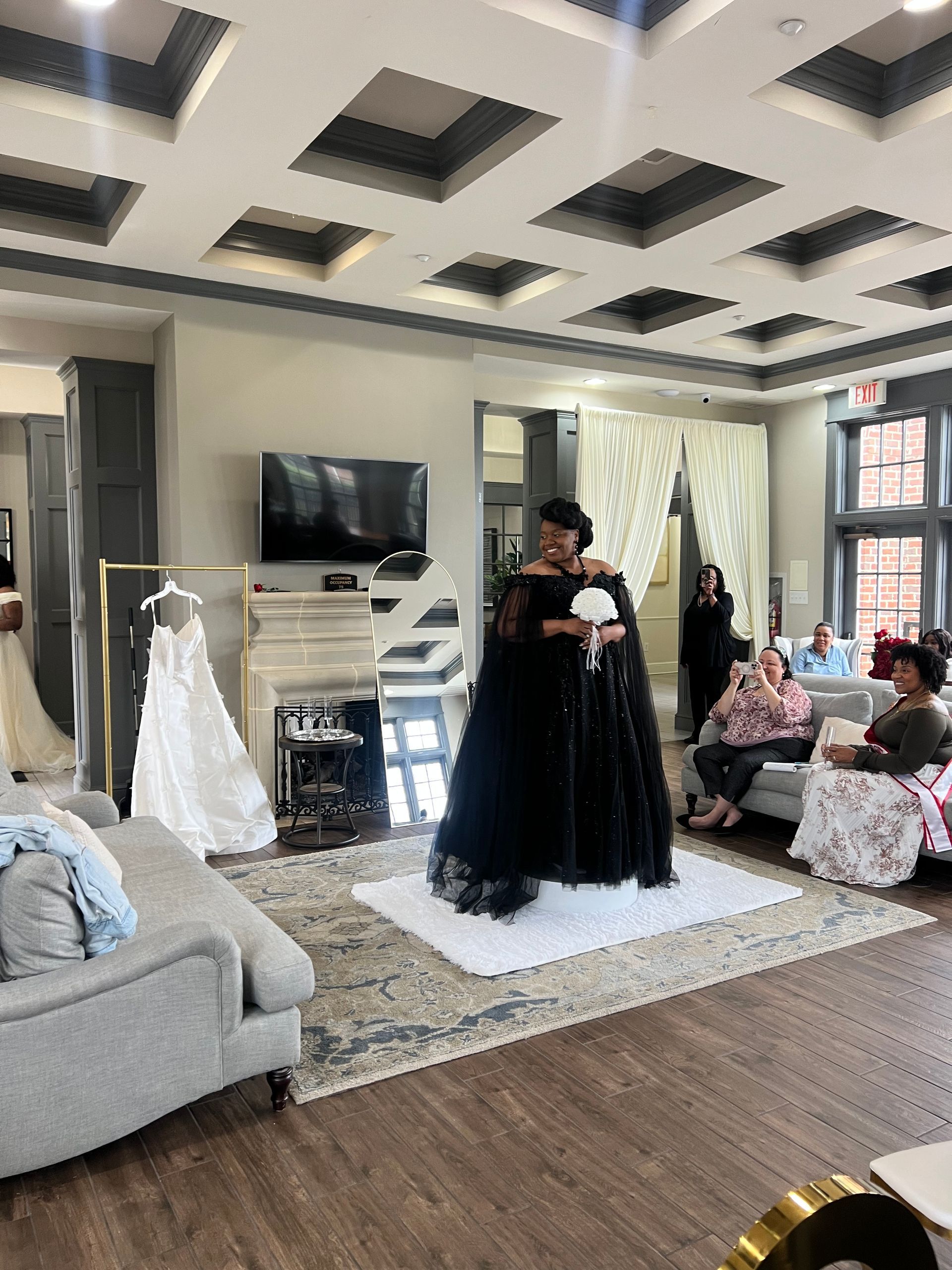 Living room bridal shower with a woman in a black gown opening a gift, surrounded by seated guests.