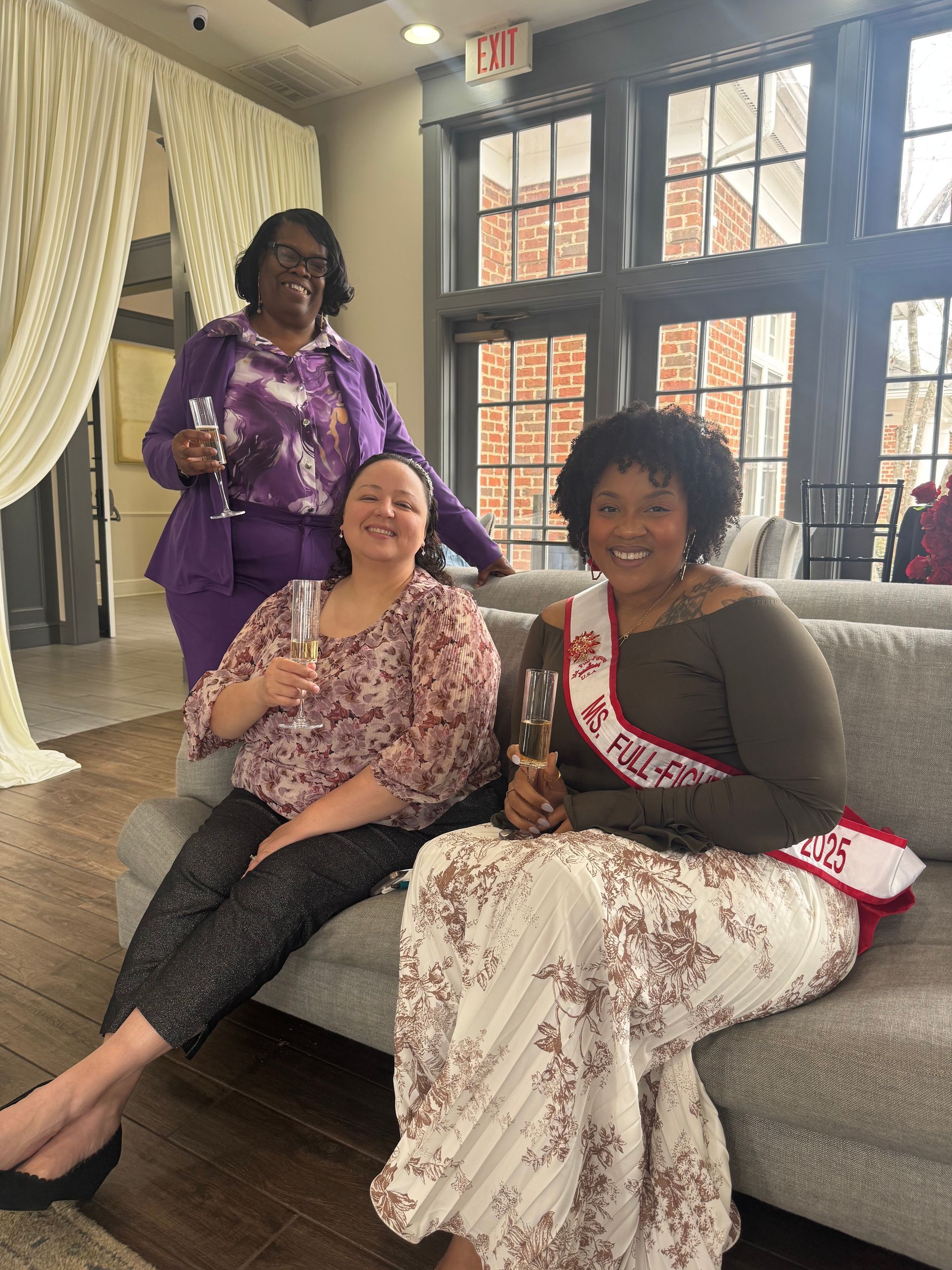Three people smiling on a couch in a bright room, one standing behind them holding a drink.