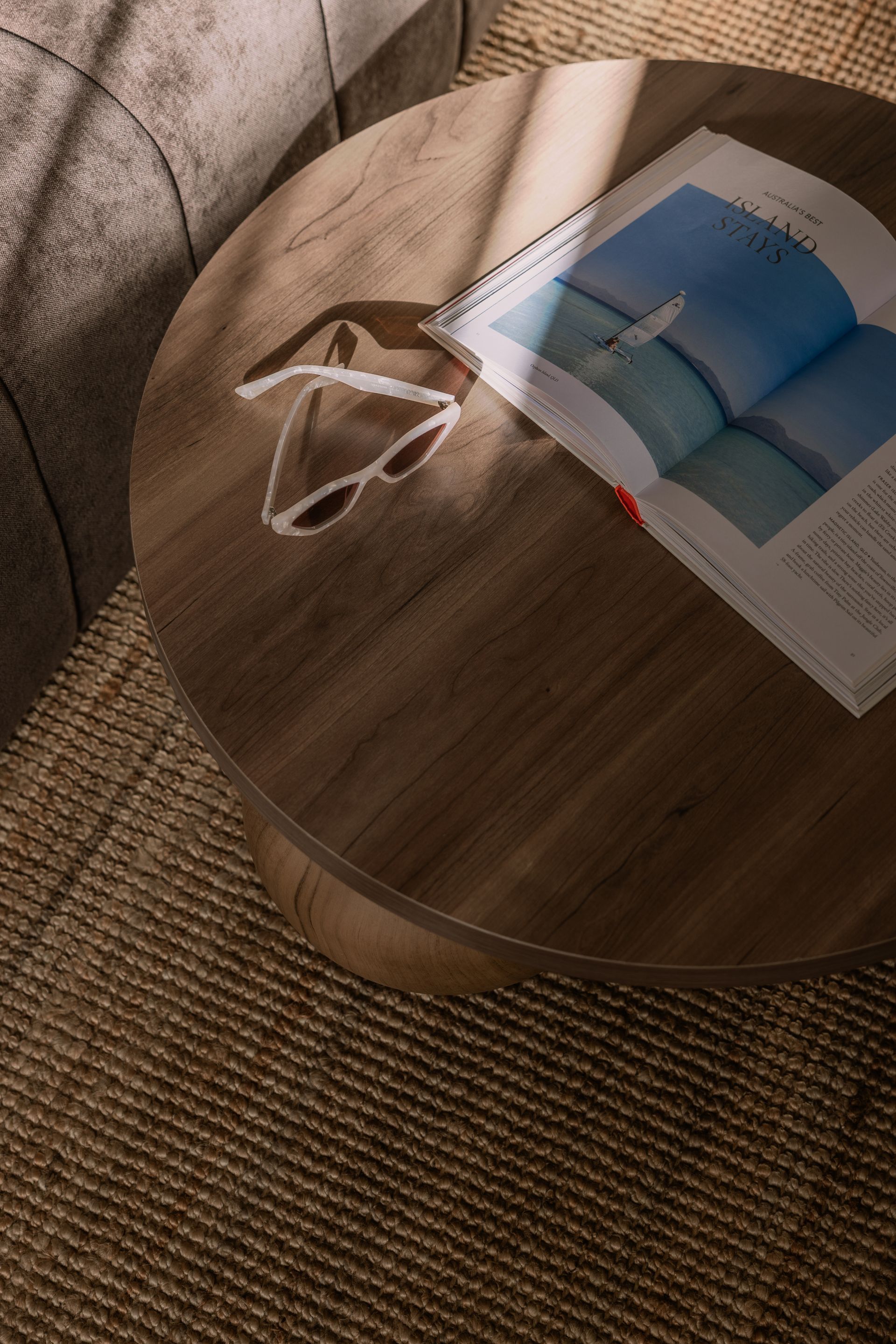 Round wooden stool with a book or magazine and white glasses on top, beside a woven rug and sofa.