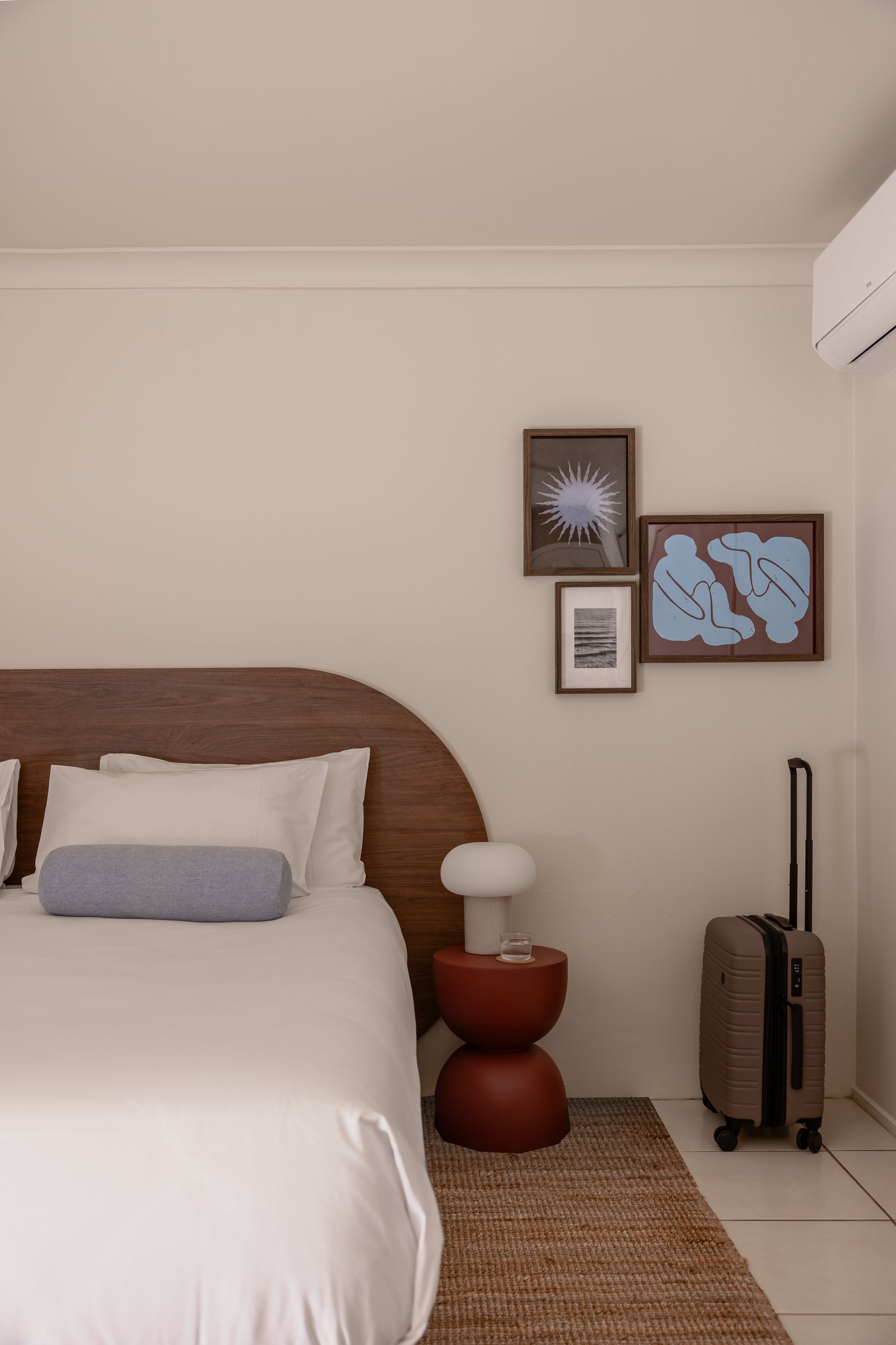 Minimal bedroom with white bed, woven rug, bedside lamp, framed art, and suitcase by the wall.