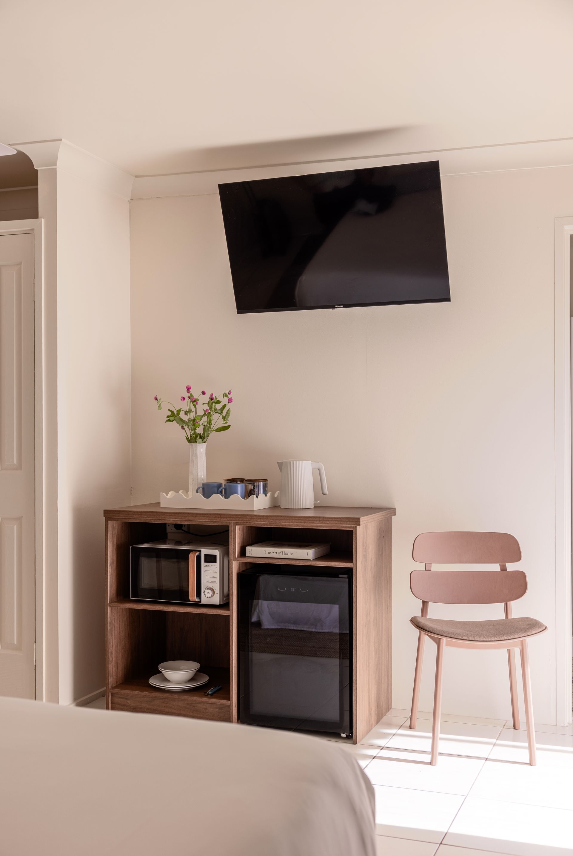 Hotel room wall with mounted TV, wooden cabinet, and pink chair near a sunlit window