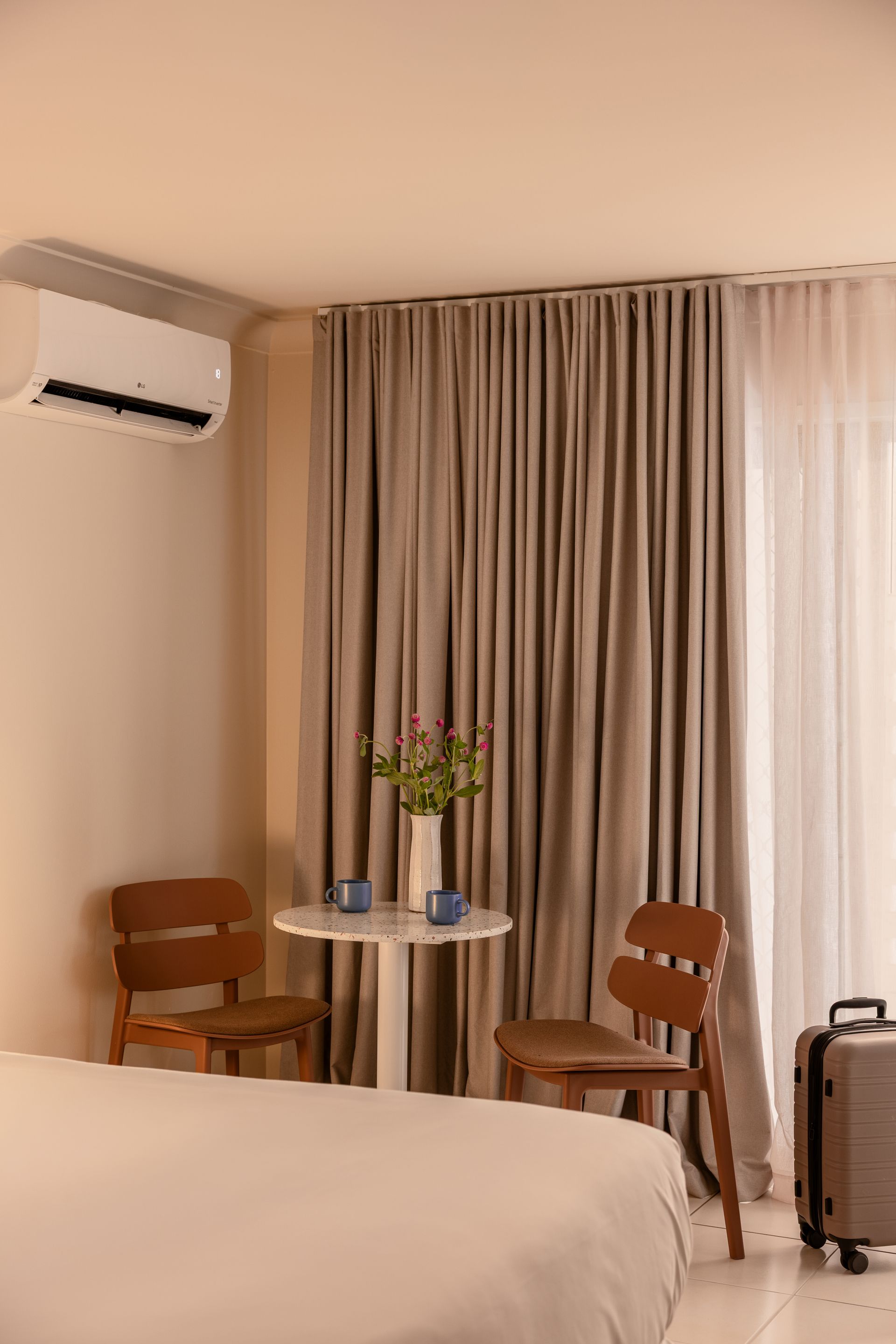 Small hotel room with bed, round table, two chairs, curtains, wall AC, and a suitcase near the window.