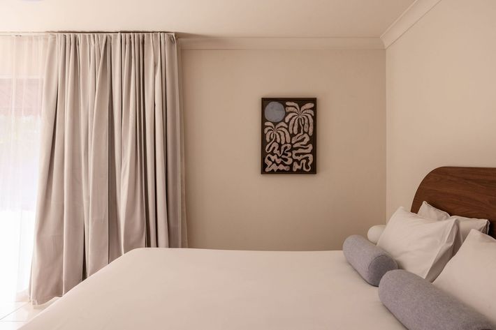 Neat beige bedroom with a bed, side tables, desk chairs, rug, and a window blind.