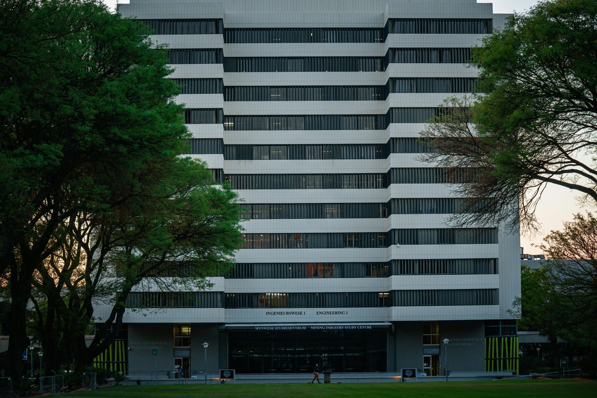 Modern white office building with rows of windows, framed by trees and a grassy foreground