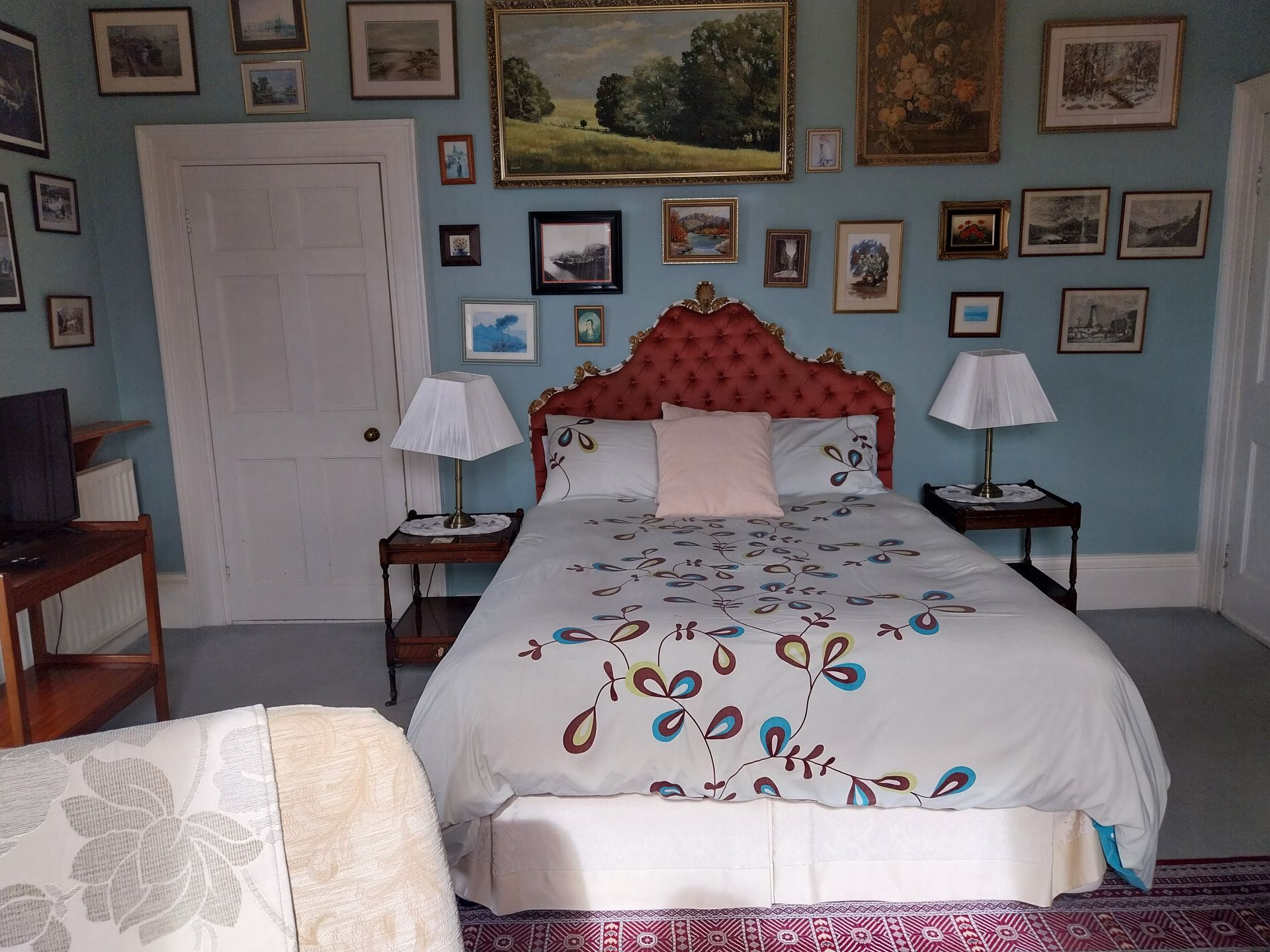 A bedroom featuring a bed with a patterned duvet and a red headboard against a blue wall covered with numerous framed photos.