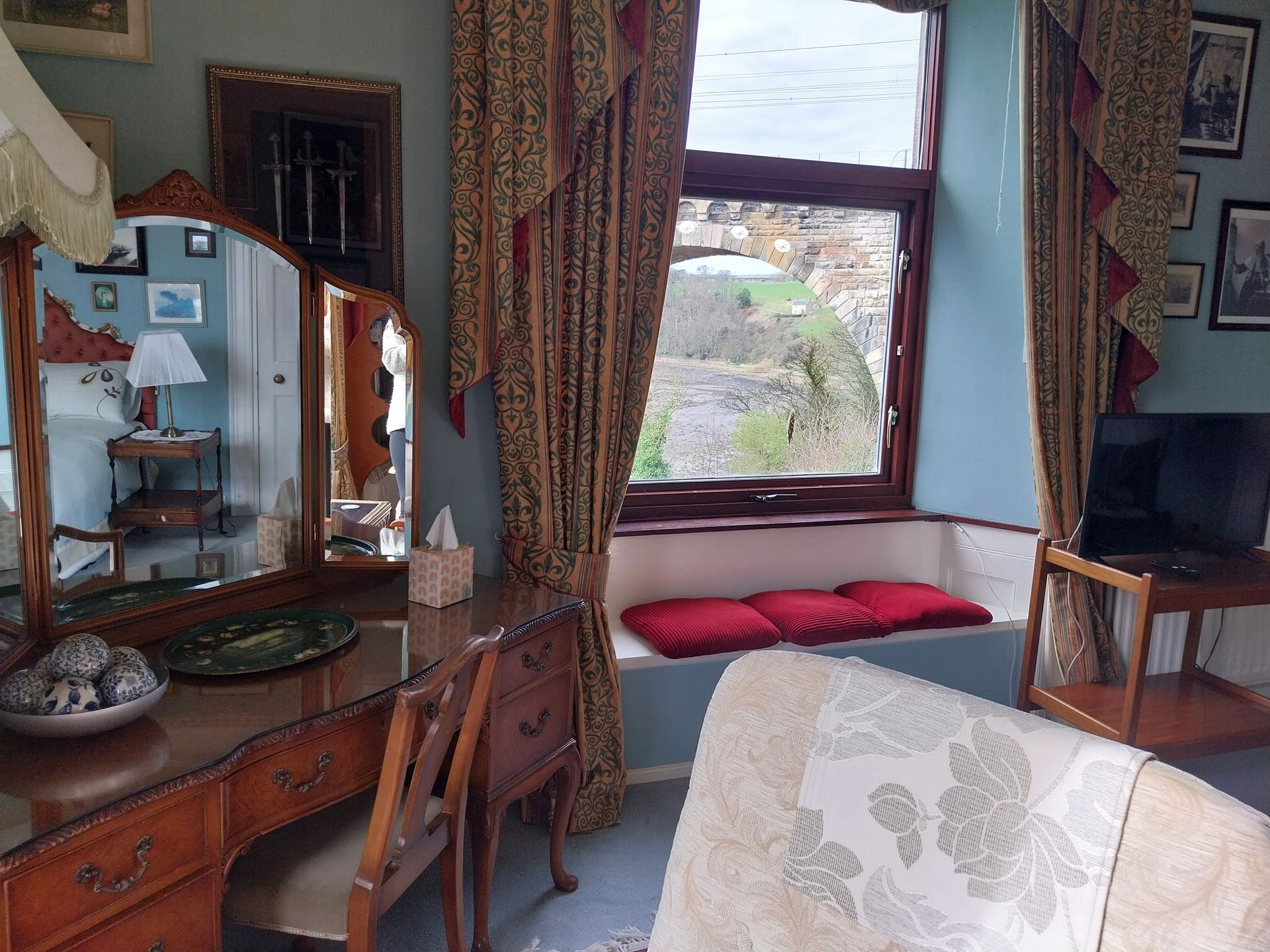 A sunlit bedroom with a wooden vanity mirror, red-cushioned window seat, patterned curtains, and a television on a stand.