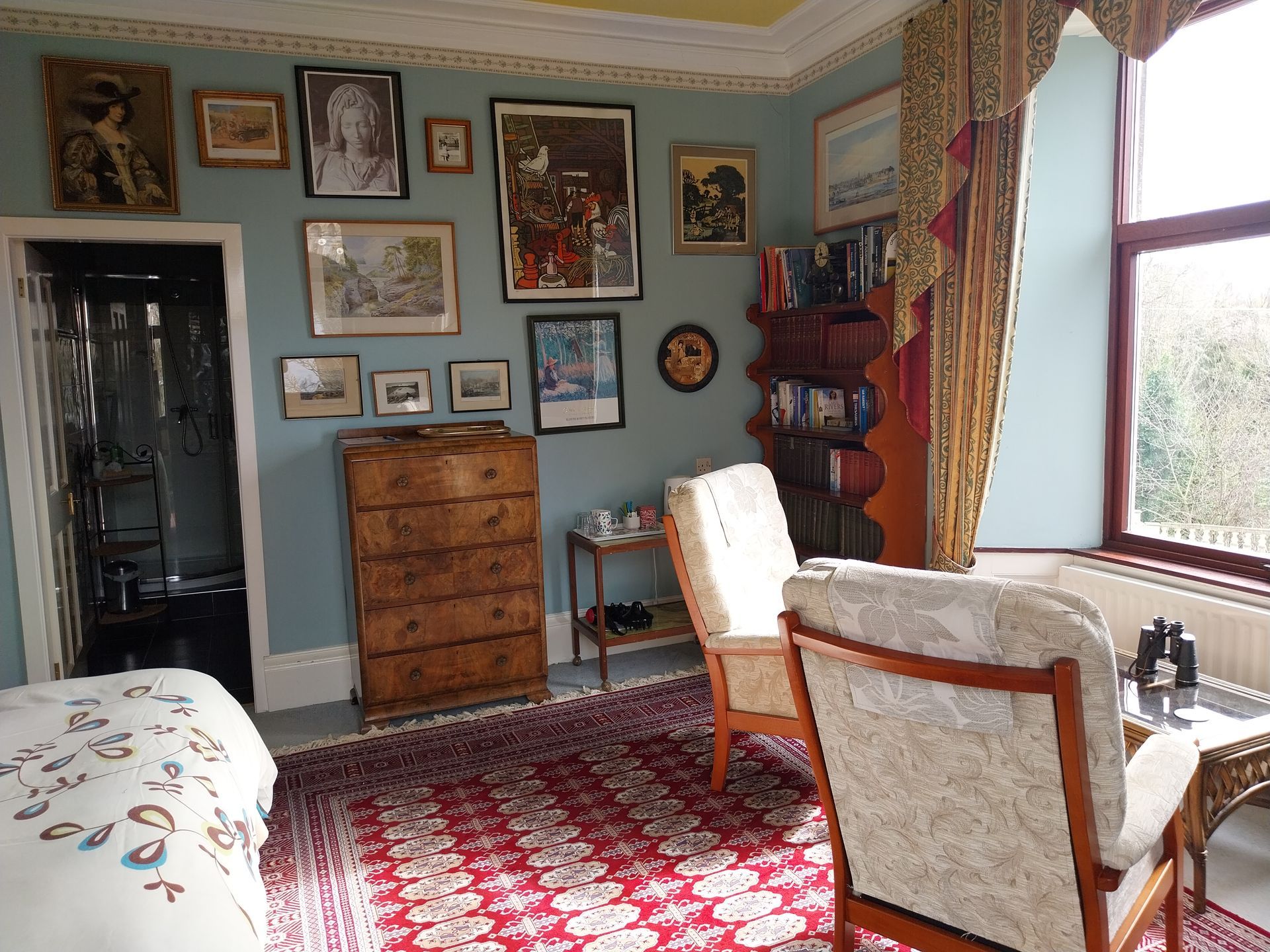 A room with blue walls decorated with many framed pictures, a wooden chest, two armchairs, and a patterned red rug.