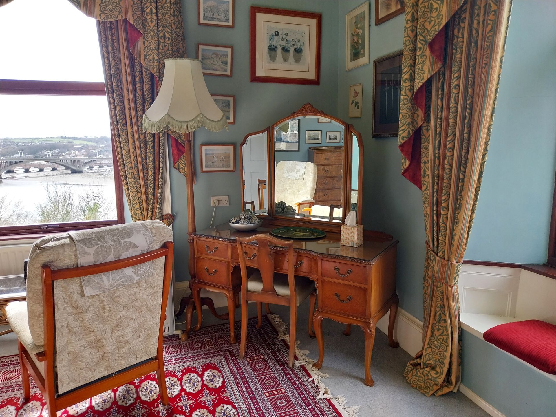 A cozy room featuring a vanity with a mirror, a patterned armchair, and a window with curtains overlooking a bridge.