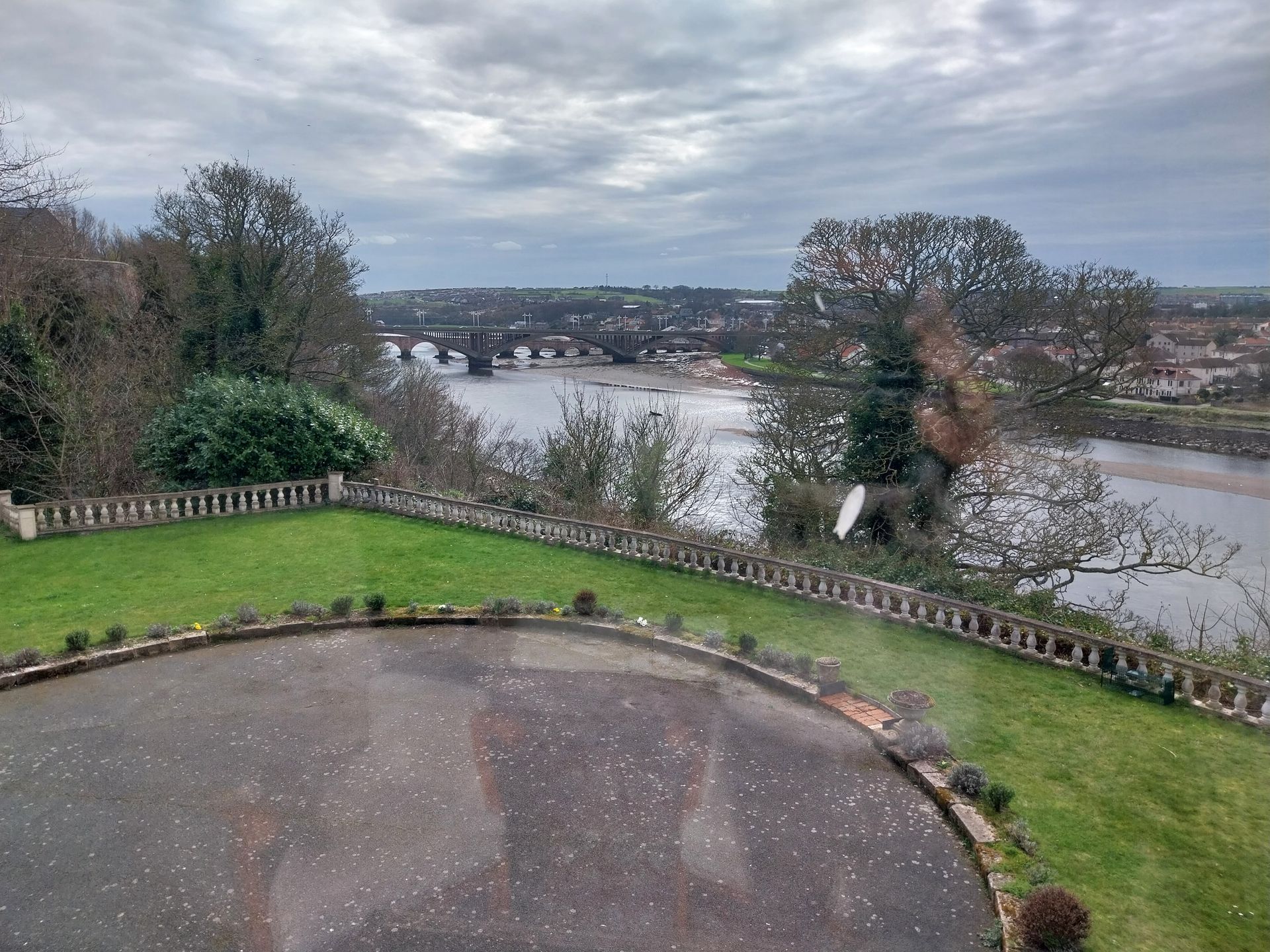 Elevated view of a paved circular driveway, green lawn, and stone fence overlooking a river with a distant arched bridge.