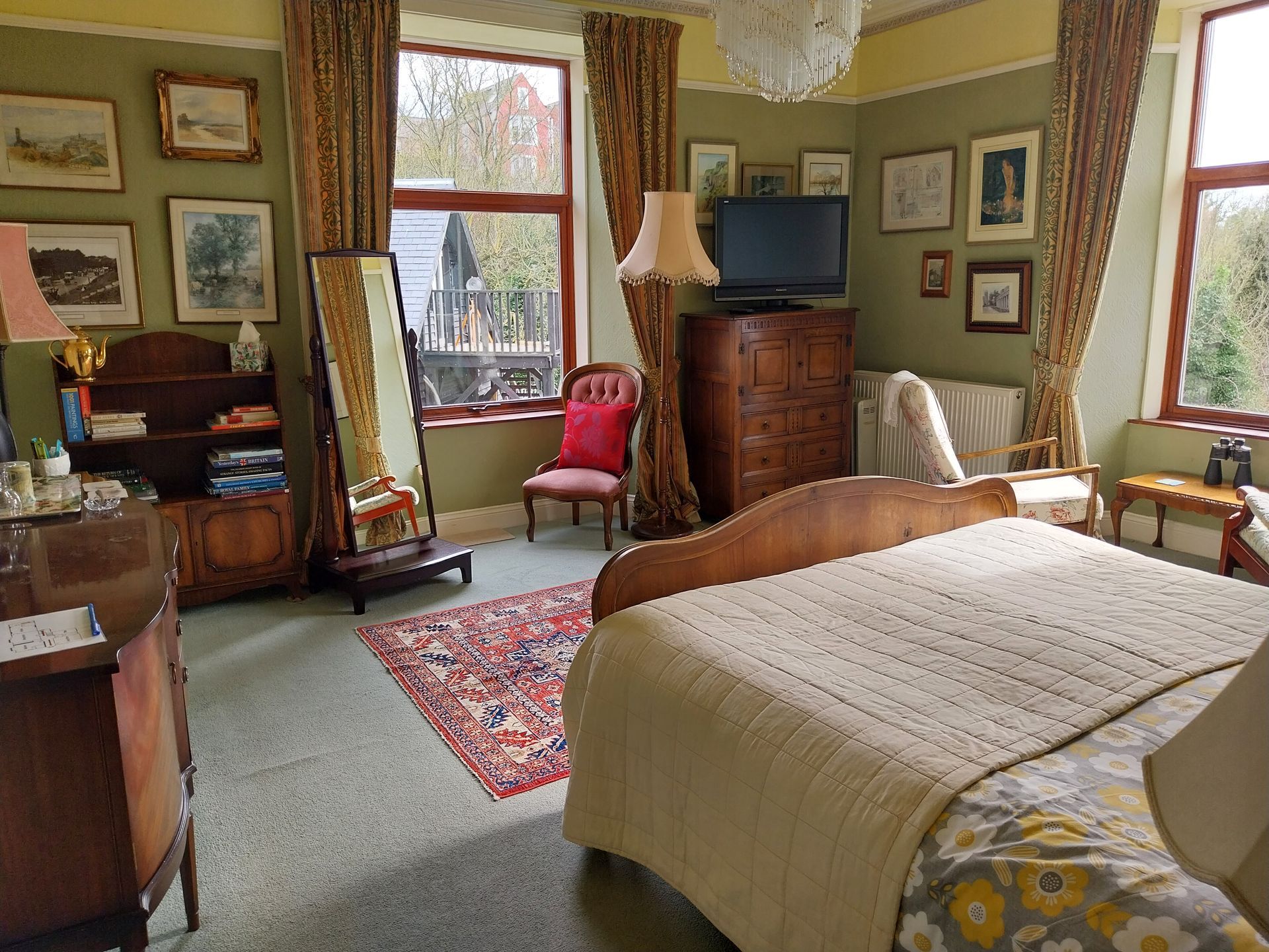 A bedroom featuring a bed, patterned rug, red armchair, wooden furniture, and framed wall art in a sage-green room.