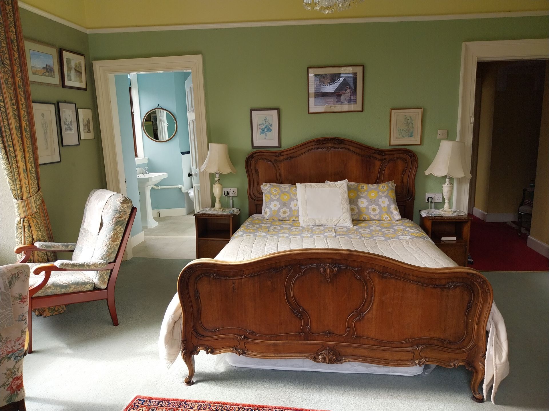A bedroom with a carved wooden bed, green walls, light-colored bedding, two lamps, and an attached bathroom entrance.