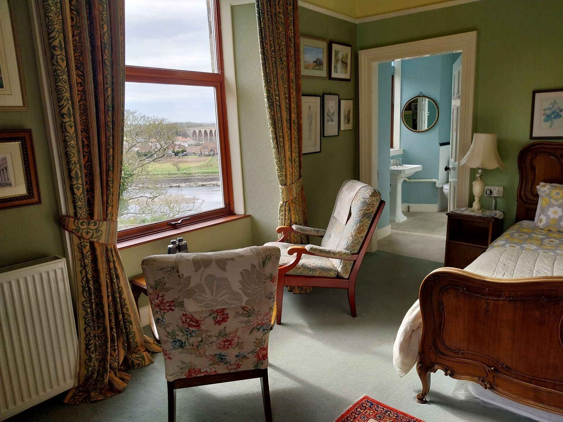 A green bedroom with a wooden bed, two upholstered chairs by a window overlooking a river, and a view into a bathroom.
