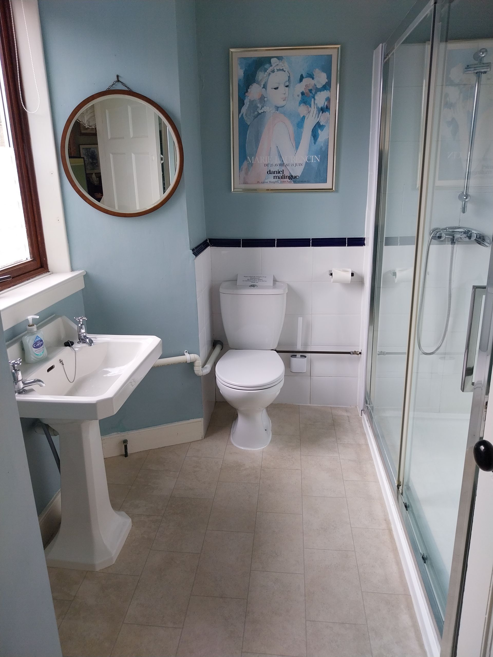 A bathroom with light blue walls, a white pedestal sink, a toilet, a round mirror, and a glass shower stall.