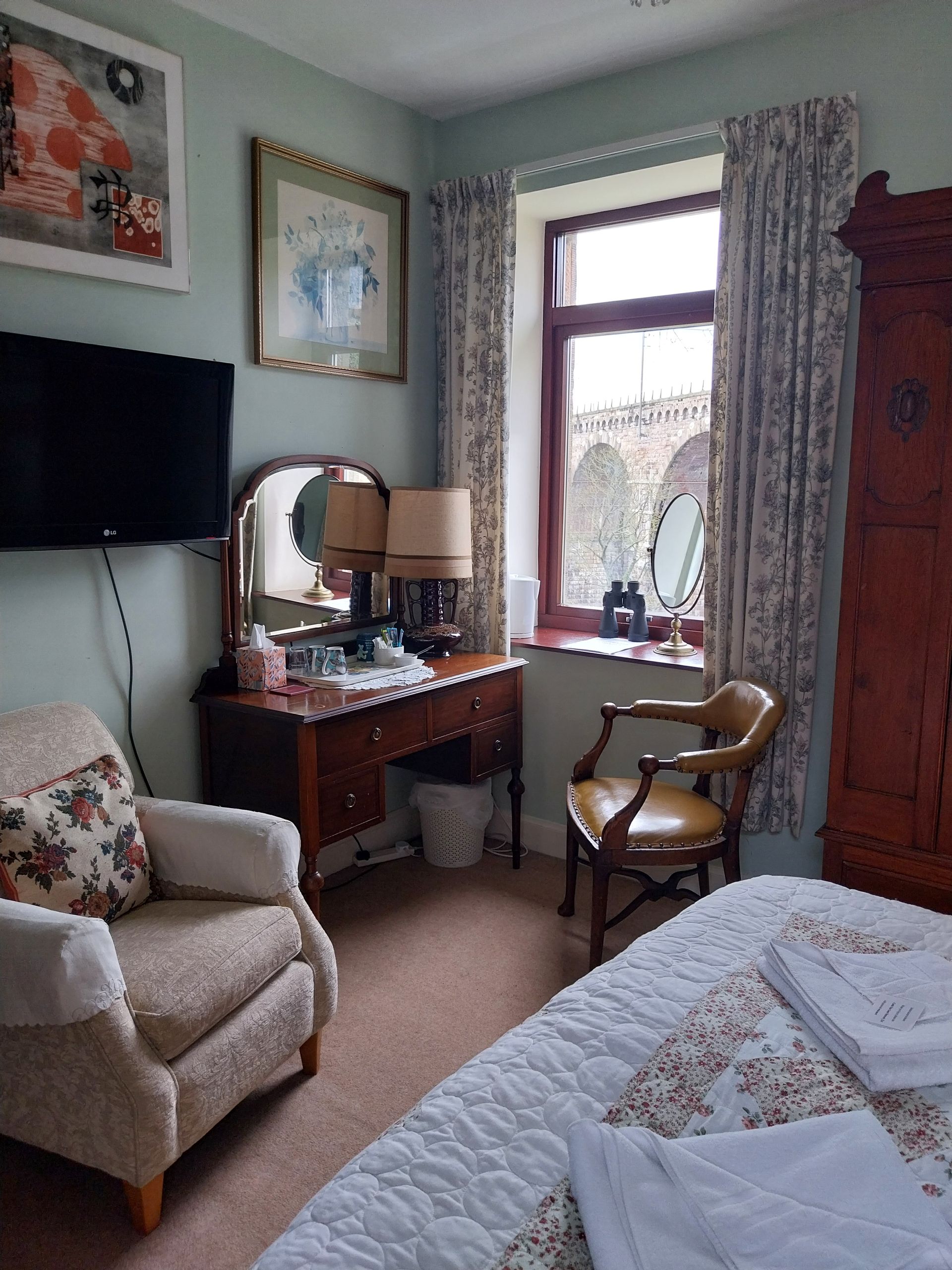 A cozy bedroom with a patterned armchair, a dark wood vanity with a mirror, a window with floral curtains, and a bed.