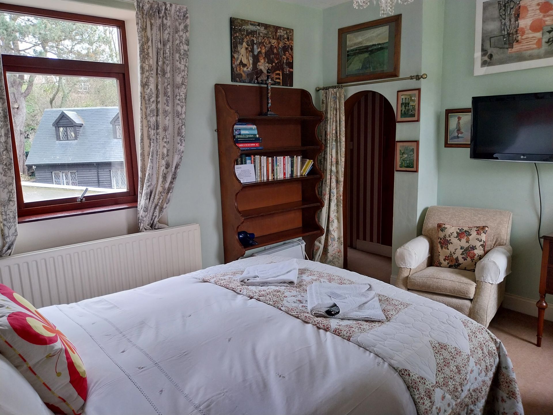 A bedroom featuring a double bed with white bedding, a wooden bookshelf, a cream armchair, and a wall-mounted TV.