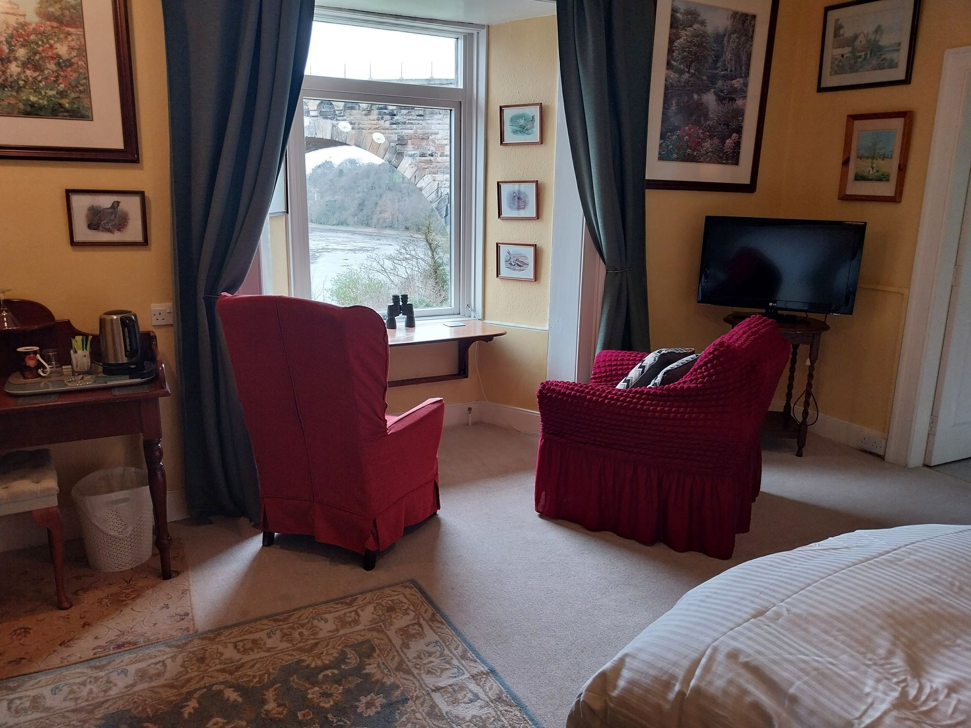 A hotel room featuring two red armchairs facing a window, a small side table, a desk, and a wall-mounted television.