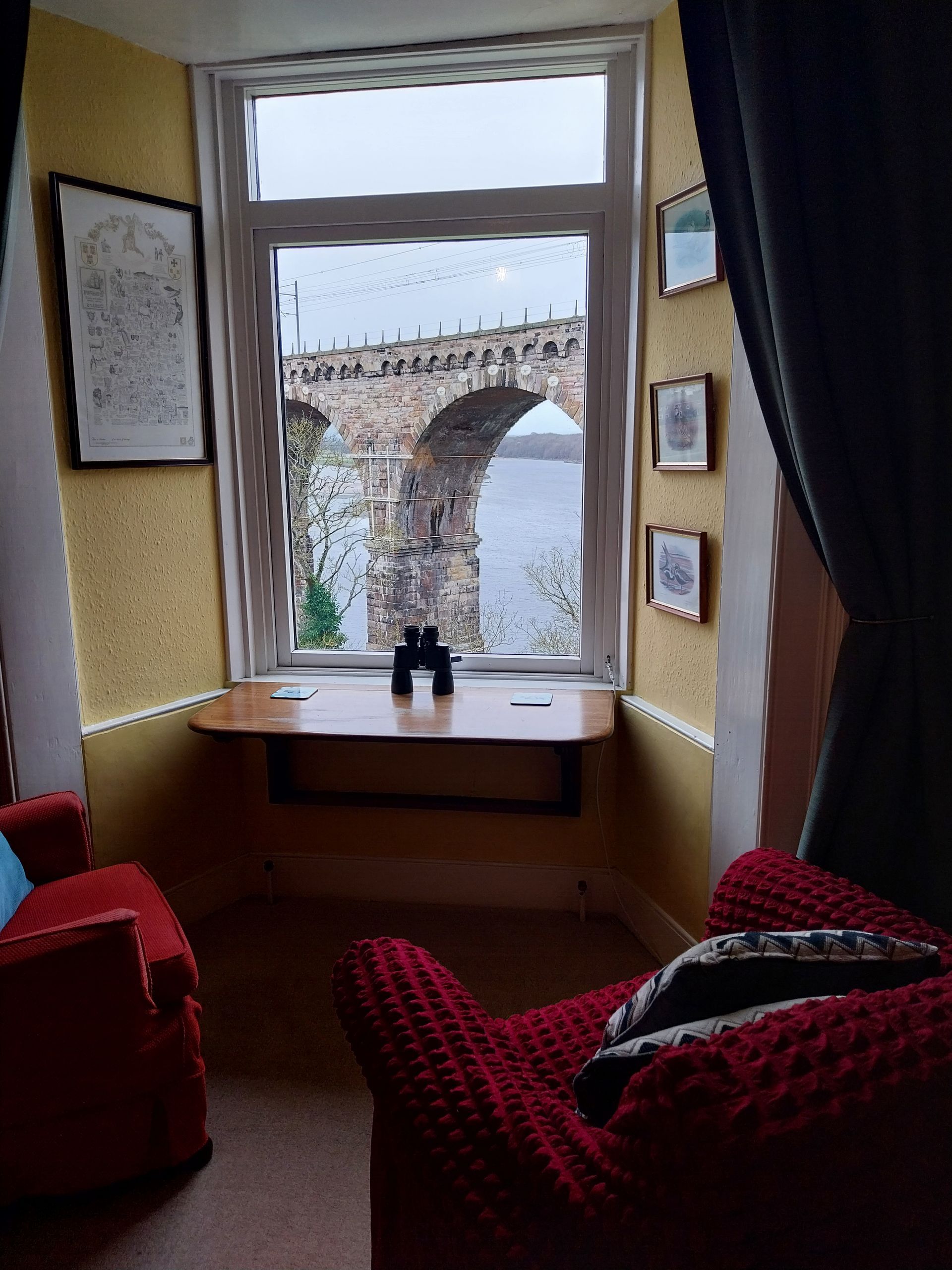 A cozy room with red chairs and a window view of an old stone bridge over water, with binoculars on a small wooden table.