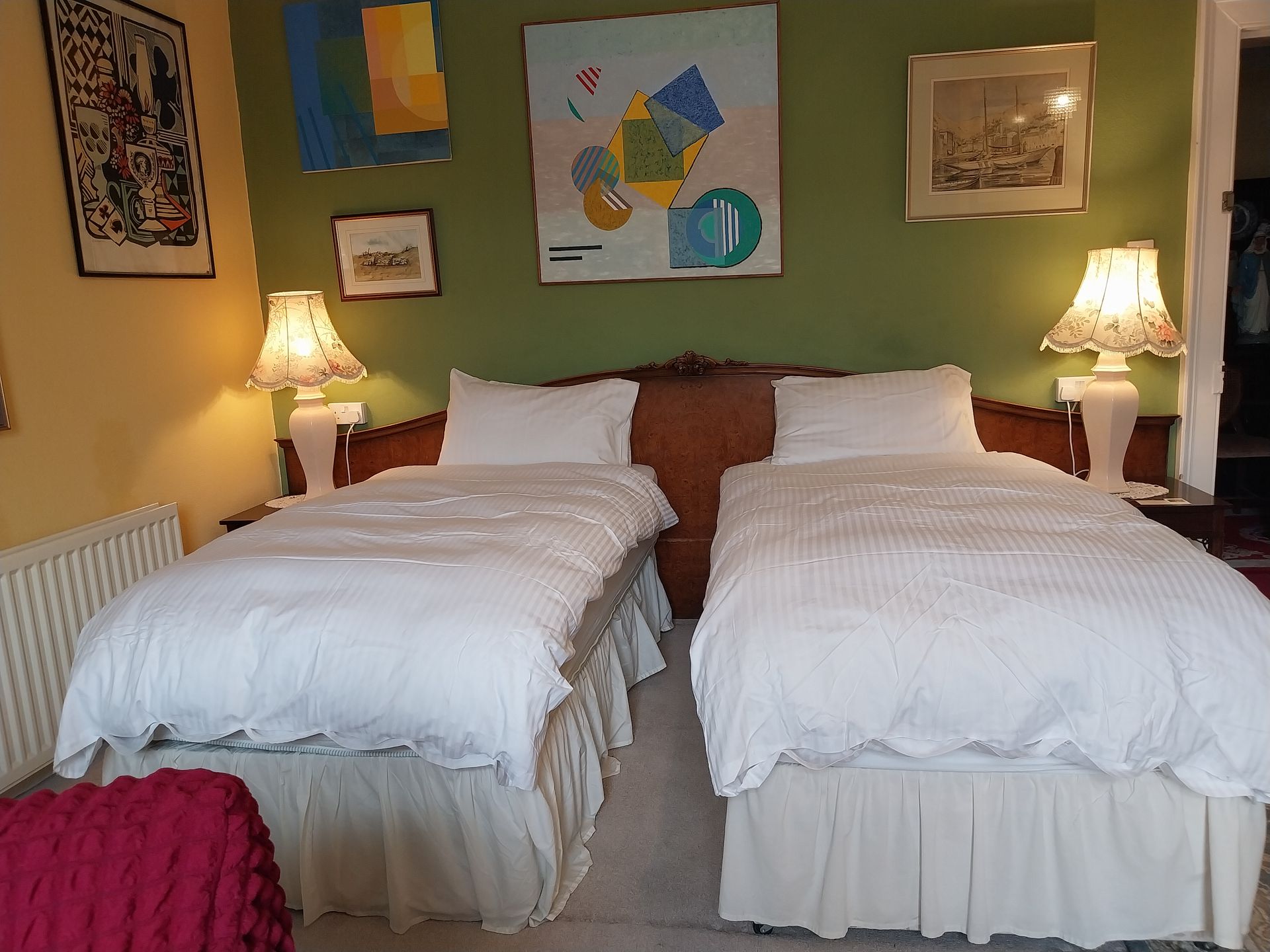 Two twin beds with white duvets and ruffled bed skirts against a green wall with three framed art pieces and two lamps.