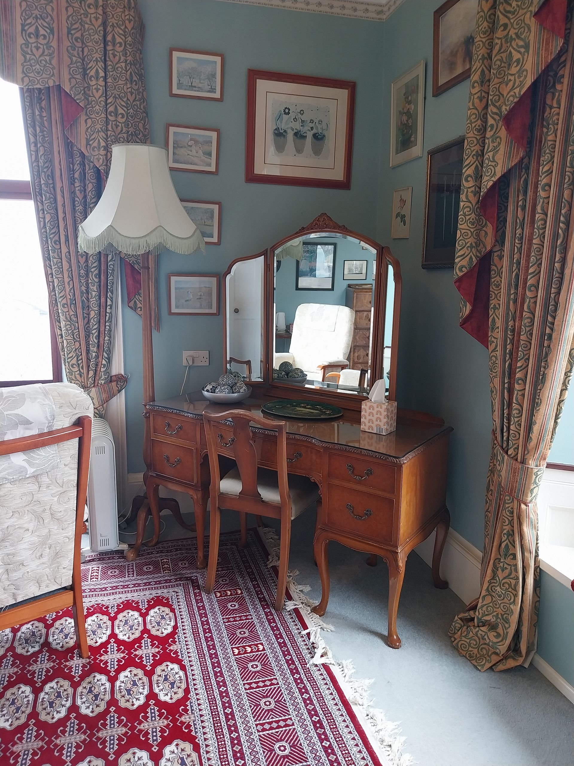 A vanity set with a tri-fold mirror and chair sits against a blue wall decorated with framed art, next to a floor lamp.