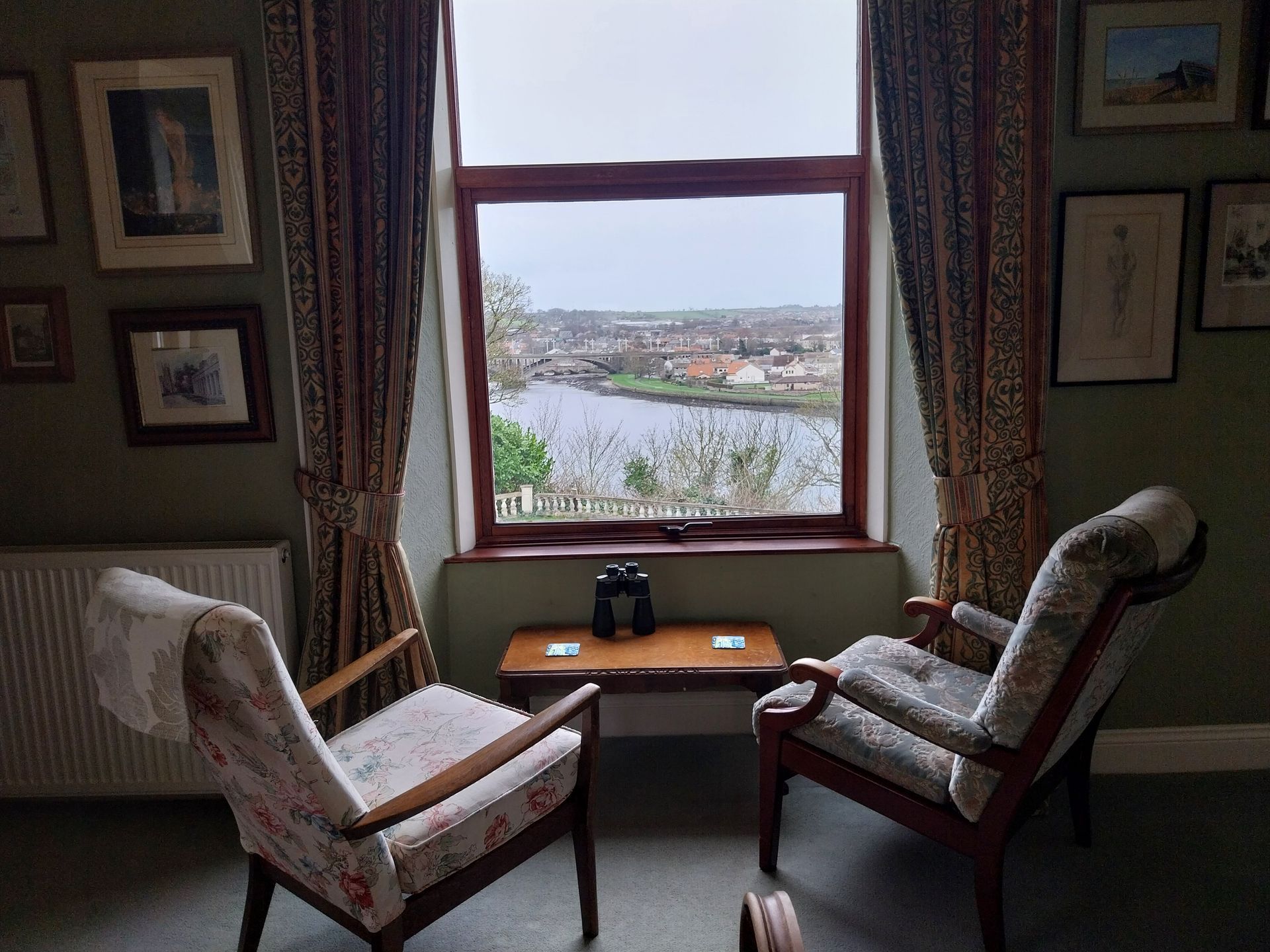Two armchairs face a window with a river view, separated by a small wooden table with binoculars in a green-walled room.