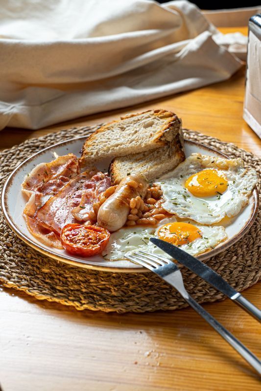 A full English breakfast with fried eggs, bacon, sausage, baked beans, a grilled tomato, and toasted bread on a plate.