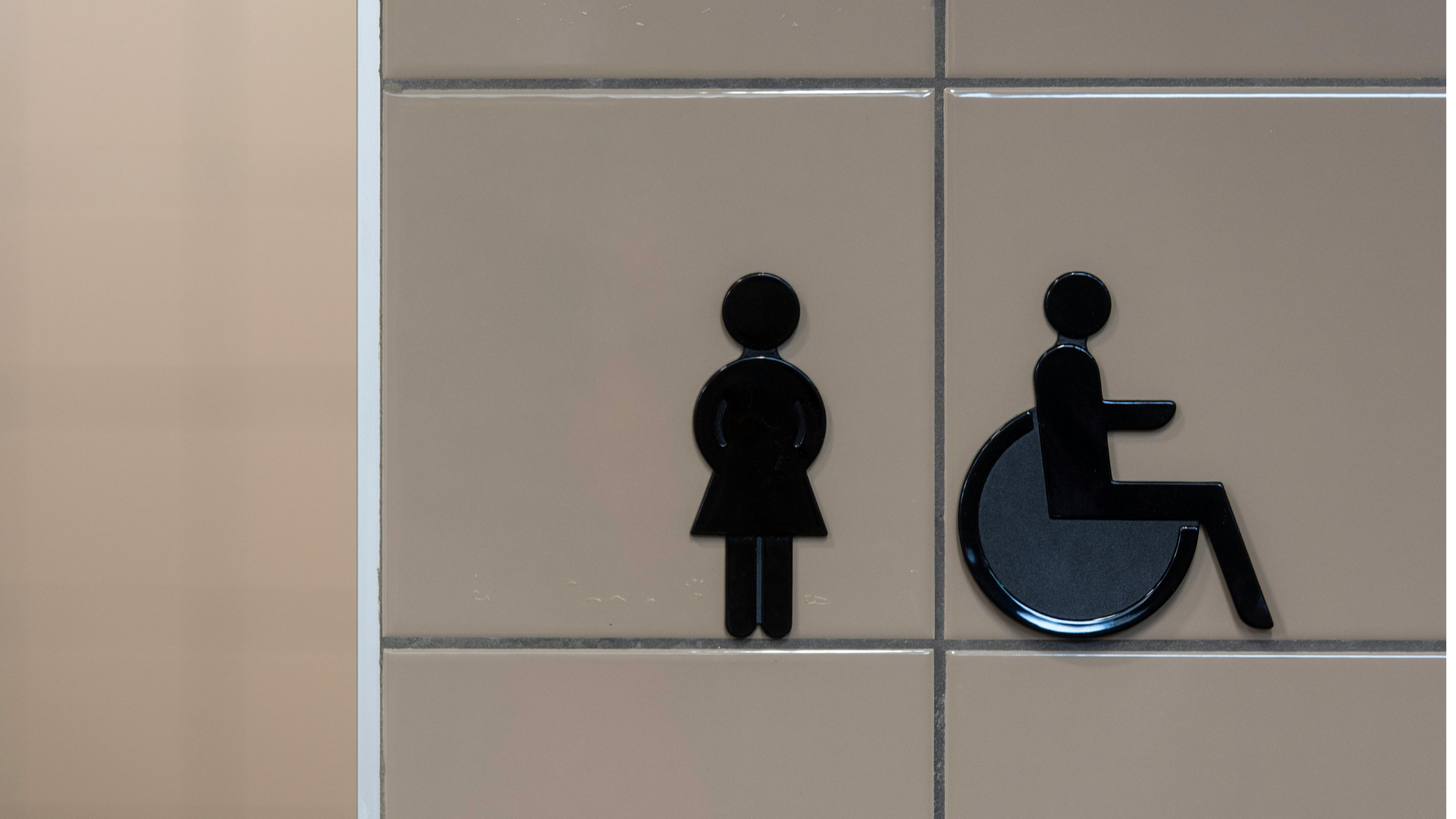Bathroom signs for women and wheelchair-accessible stall on beige tiled wall.