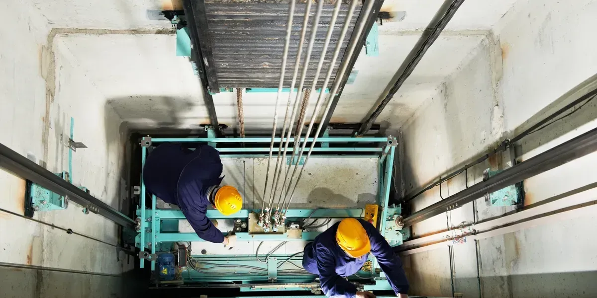 Blue elevator platform inside a shaft. Tools and control panel visible. Doorway on the right.