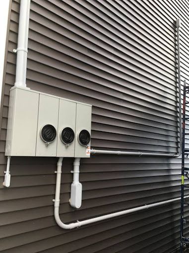 Exterior utility boxes and pipes mounted on a beige wall beside dark horizontal siding