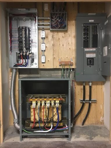 Electrical panel room with exposed wiring, breakers, and utility boxes mounted on a wooden wall.