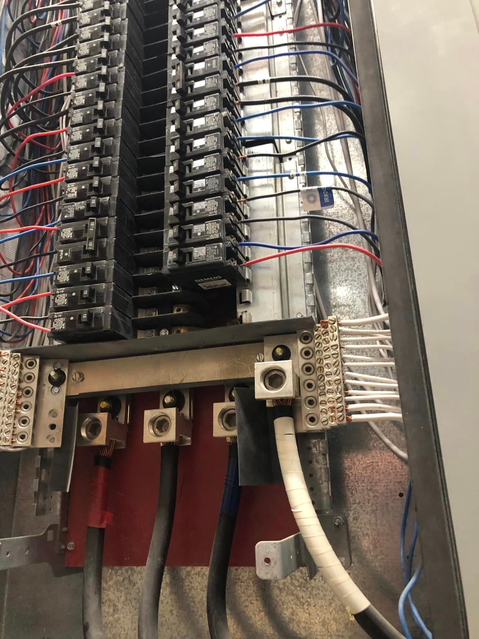 Electrical breaker panel with rows of black breakers, red wires, and white conduit inside a metal cabinet.