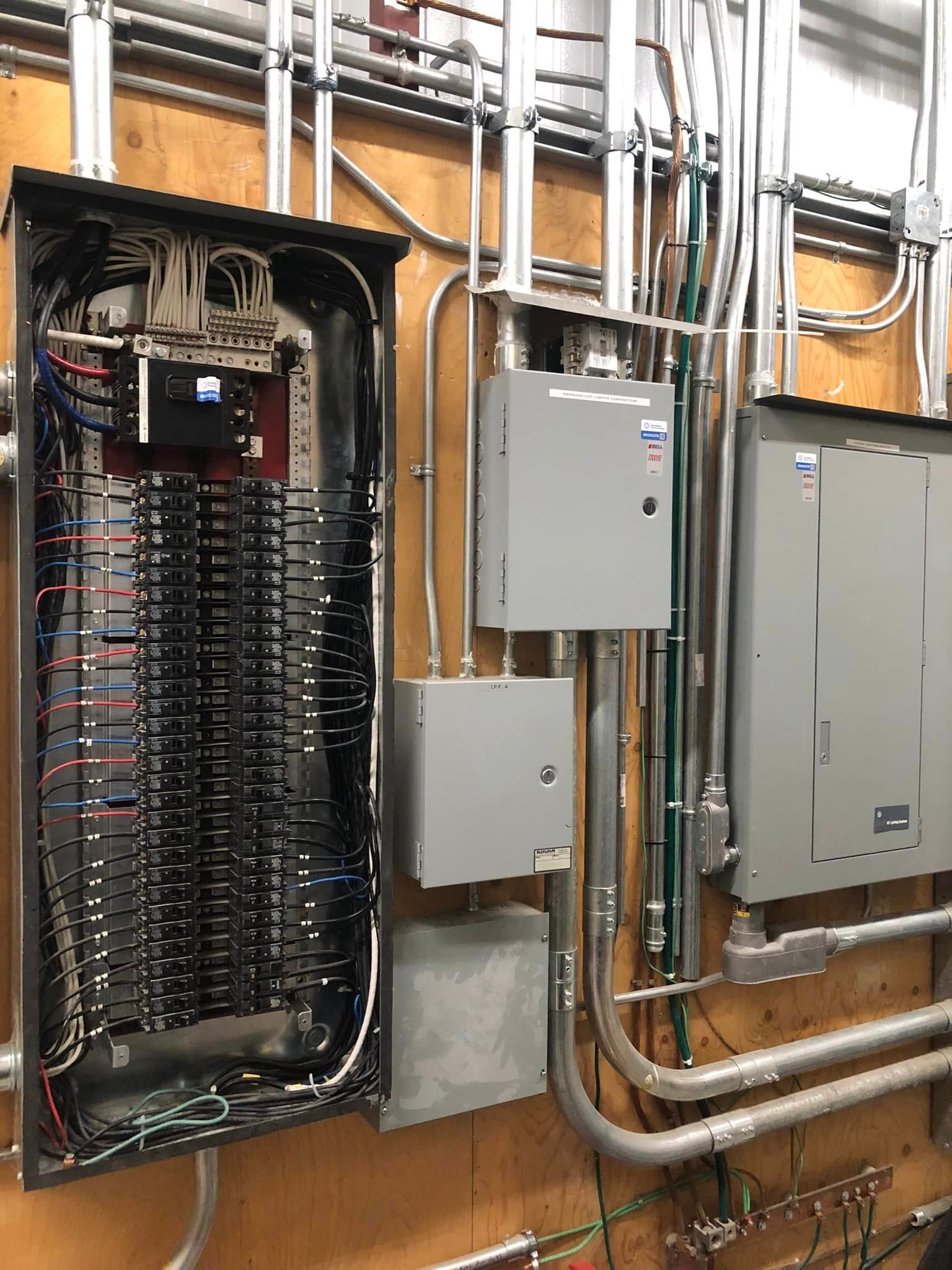 Open electrical panels with circuit breakers and conduit on a utility room wall.