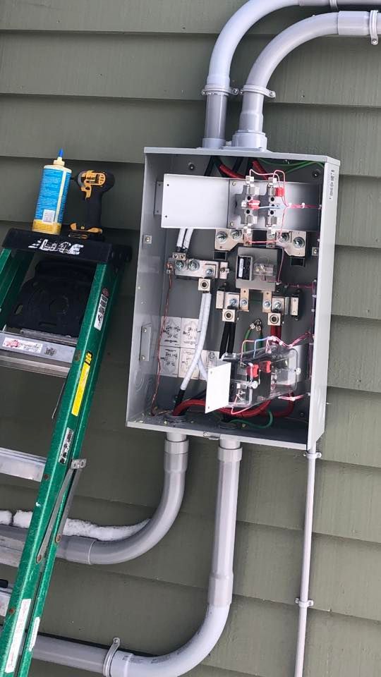 Open electrical panel mounted on green siding, with conduit pipes and tools nearby.