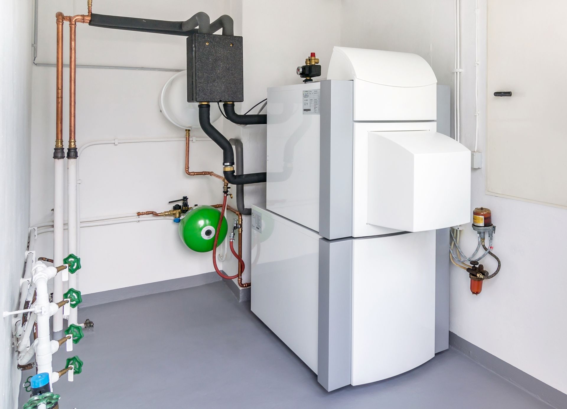 Utility room with white boiler, green expansion tank, and exposed pipes on a gray floor