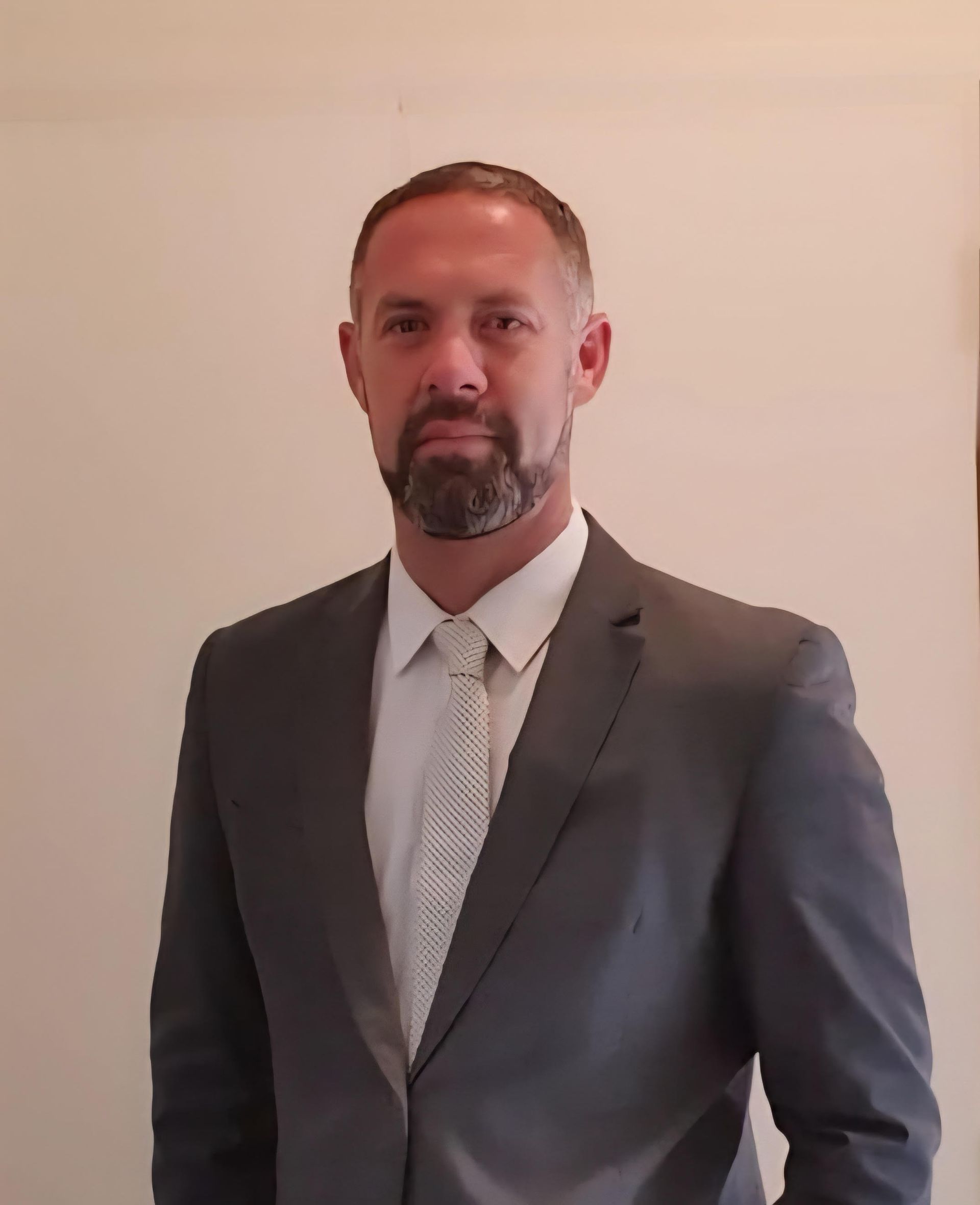 Man in a gray suit, white shirt, and patterned tie, standing in front of a neutral wall, looking forward.