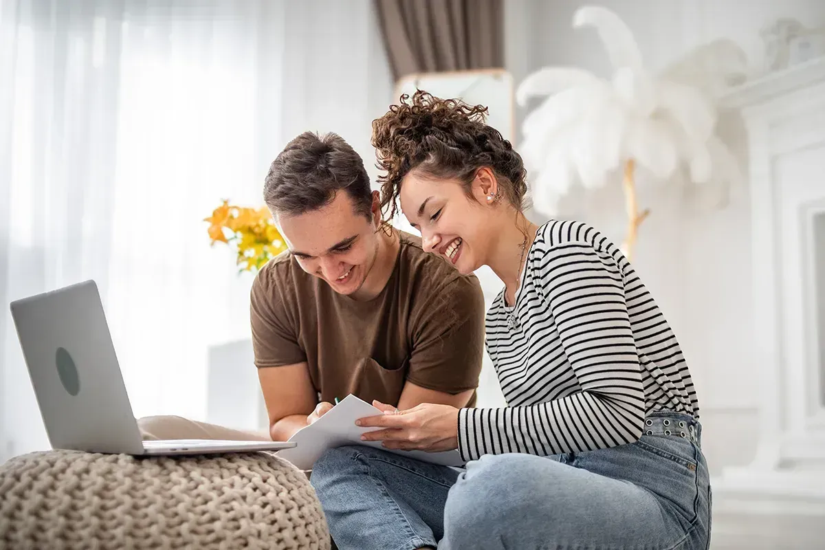Couple smiling, reviewing paperwork together near laptop in bright living room.