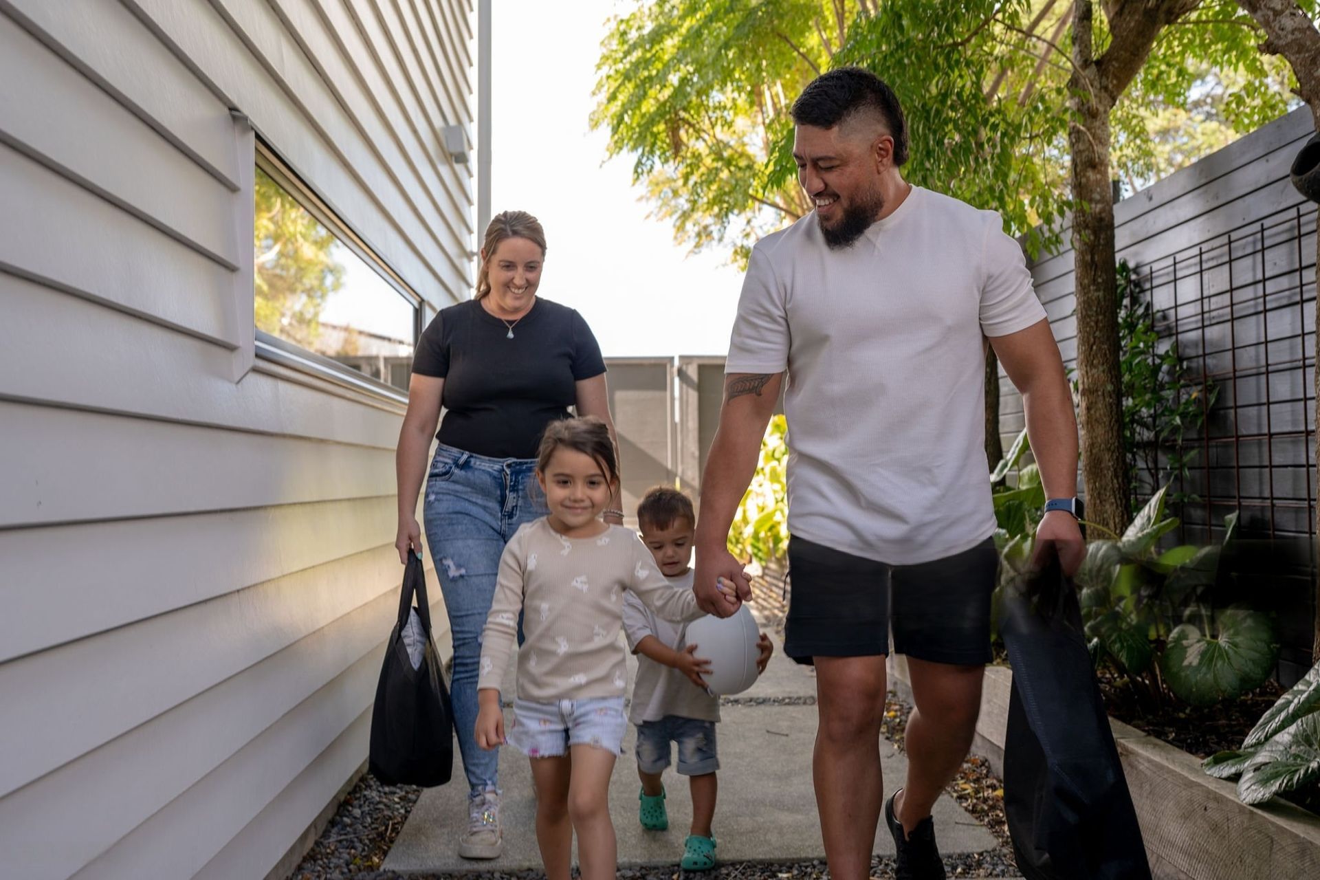 Family of four walking down a pathway; parents holding bags, children holding hands and a ball, near a building.