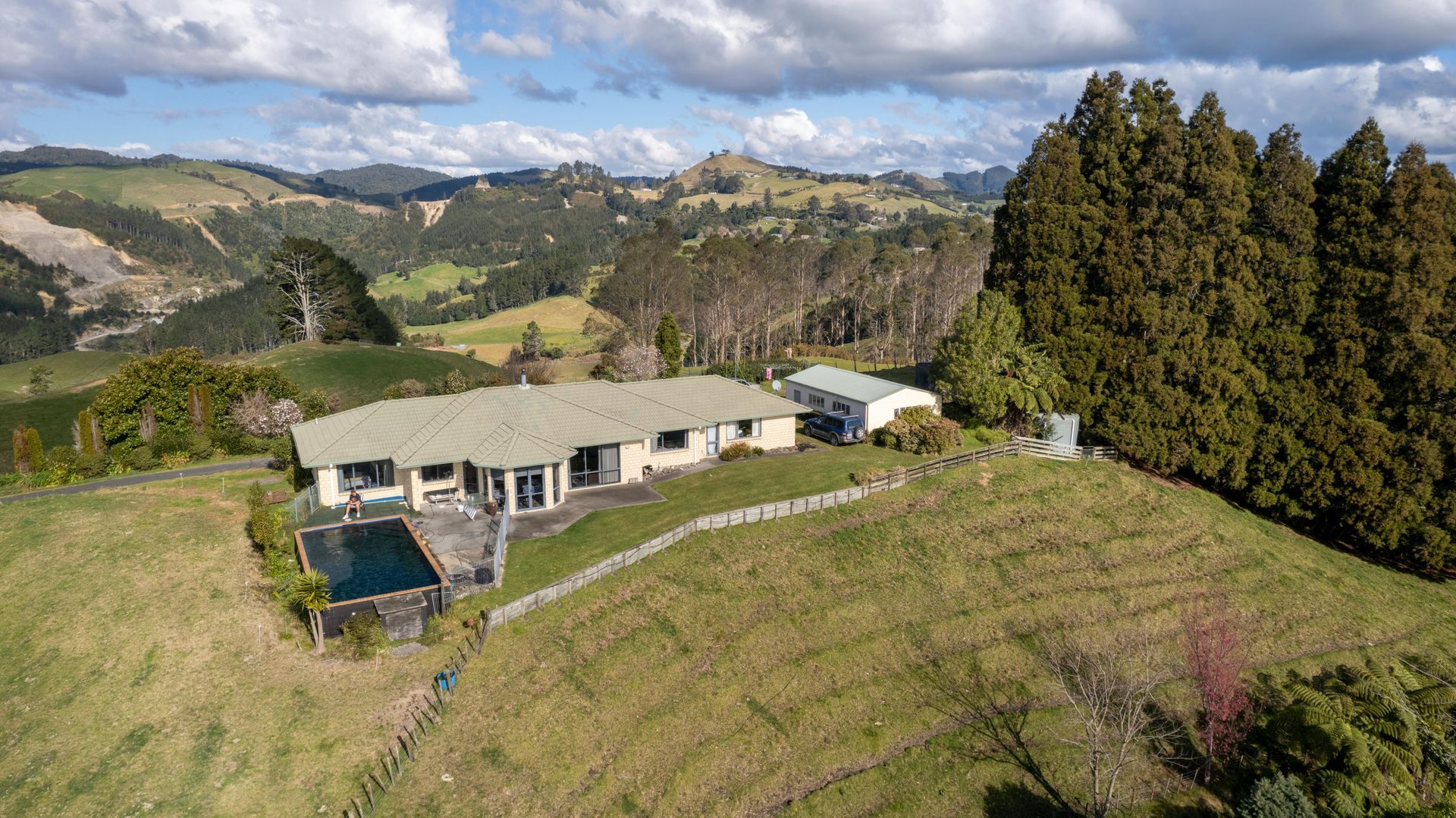 House on a grassy hill with a pool, overlooking rolling hills and trees under a cloudy sky.